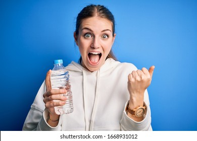 Young beautiful redhead woman doing sport drinking bottle of water over blue background screaming proud and celebrating victory and success very excited, cheering emotion - Shutterstock ID 1668931012