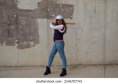 young and beautiful redhead woman is happy with baseball cap, jacket and baseball bat in position to hit the ball. it is on grey cement background. Concept sport and recreations.