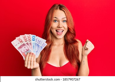Young beautiful redhead woman holding swedish krona banknotes screaming proud, celebrating victory and success very excited with raised arms 
