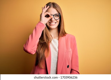 Young beautiful redhead woman wearing jacket and glasses over isolated yellow background doing ok gesture with hand smiling, eye looking through fingers with happy face.