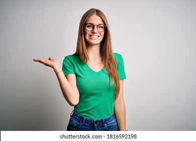 Young beautiful redhead woman wearing casual green t-shirt and glasses over white background smiling cheerful presenting and pointing with palm of hand looking at the camera.