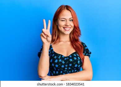 Young beautiful redhead woman wearing casual clothes over blue background smiling with happy face winking at the camera doing victory sign with fingers. number two.  - Powered by Shutterstock