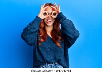 Young Beautiful Redhead Woman Wearing Casual Sweater And Glasses Over Blue Background Doing Ok Gesture Like Binoculars Sticking Tongue Out, Eyes Looking Through Fingers. Crazy Expression. 