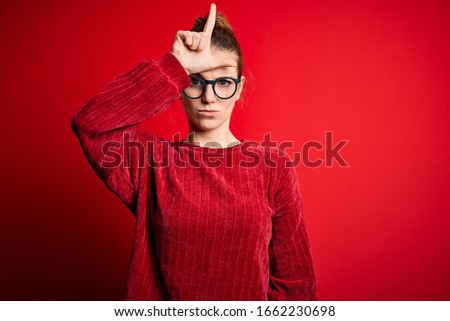 Young beautiful redhead woman wearing casual sweater over isolated red background making fun of people with fingers on forehead doing loser gesture mocking and insulting.