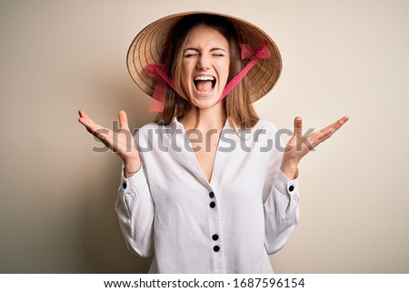Young beautiful redhead woman wearing asian traditional conical hat over white background celebrating mad and crazy for success with arms raised and closed eyes screaming excited. Winner concept
