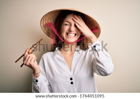 Young beautiful redhead woman wearing asian traditional hat holding wooden chopsticks stressed with hand on head, shocked with shame and surprise face, angry and frustrated. Fear and upset for mistake