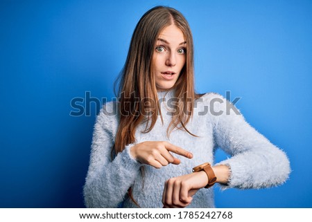 Young beautiful redhead woman wearing casual sweater over isolated blue background In hurry pointing to watch time, impatience, upset and angry for deadline delay