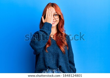 Young beautiful redhead woman wearing casual sweater and glasses over blue background covering one eye with hand, confident smile on face and surprise emotion. 