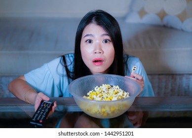 Young Beautiful And Scared Asian Chinese Teenager Woman In Fear Watching Horror Scary Movie At Home Sofa Couch Eating Popcorn Bowl Holding Remote Controller In Horrified Face Expression