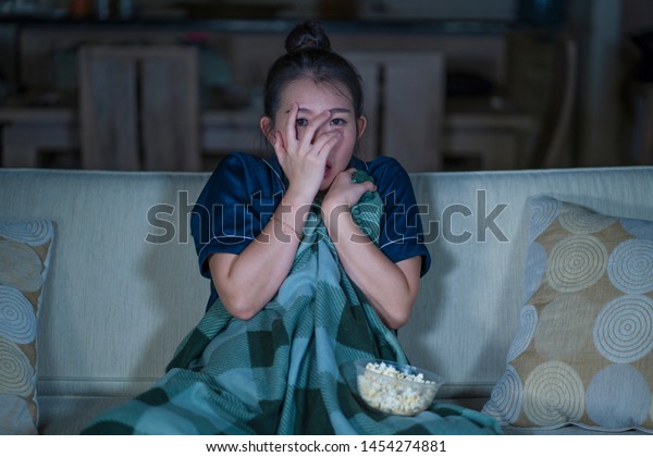  young beautiful scared and frightened Asian\
Japanese woman watching horror scary movie or thriller eating\
popcorn in fear face expression eating popcorn sitting at living\
room couch in the dark