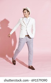 Young Beautiful Serious Modern Man Wearing White Jacket, White Shirt, Grey Pants And White Hat Stepping Through Studio With Light Pink Background