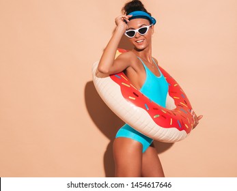 Young beautiful sexy smiling hipster woman in sunglasses.Girl in summer swimwear bathing suit with donut lilo inflatable mattress.Positive female going crazy.Near beige wall in transparent visor cap
