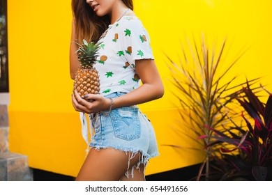 young beautiful sexy stylish woman holding pineapple in hand on yellow background, summer style, positive, hips, booty, diet, denim jeans shorts