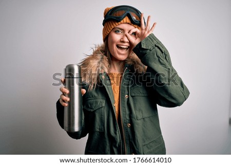 Young beautiful skier woman with blue eyes wearing ski goggles drinking thermo with coffee with happy face smiling doing ok sign with hand on eye looking through fingers