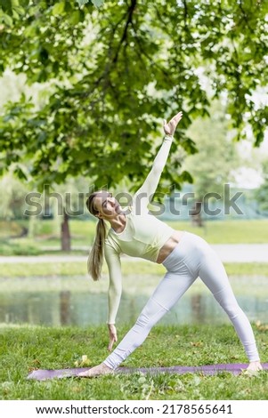 A young beautiful slender girl with long blonde hair does yoga in the summer in nature by the pond in the park.