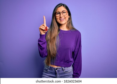 Young beautiful smart woman wearing glasses over purple isolated background showing and pointing up with finger number one while smiling confident and happy.