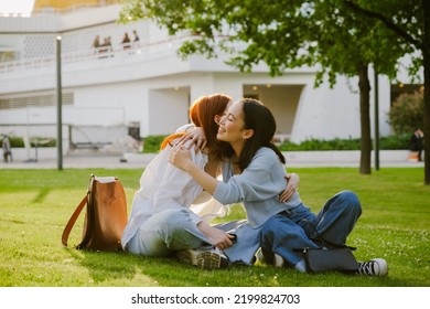 Young beautiful smiling asian girl with closed eyes hugging her redhead friend , while they sitting on the grass - Powered by Shutterstock