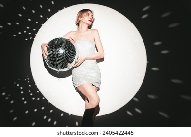 Young beautiful smiling blond female in trendy evening silver dress. Sexy carefree woman posing near white wall in studio in a circle of light. Fashionable model with bright makeup. Holds disco ball: zdjęcie stockowe