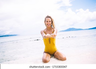 Young beautiful smiling blonde in yellow swimsuit sitting and showing seashell in hands on beach against background of sea and mountains
