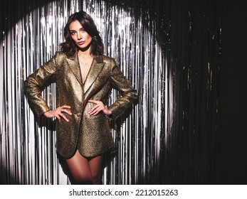 Young beautiful smiling brunette female in trendy evening golden jacket. carefree woman posing near silver shiny tinsel wall in studio. Fashionable model with bright makeup looks at camera