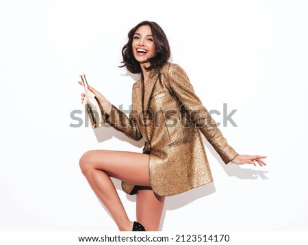 Young beautiful smiling brunette female in trendy evening golden jacket.Sexy carefree woman posing near white wall in studio.Fashionable model with bright makeup holding champagne bottle
