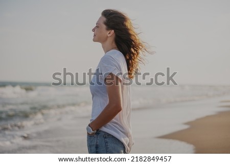 Young beautiful smiling cheerful woman in white t-shirt standing on the seashore enjoying nature