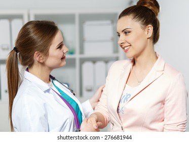 Young Beautiful Smiling Female Doctor Shaking Hand With Pretty Woman Patient. Partnership, Trust And Medical Ethics Concept. Handshake With Satisfied Client.