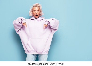 Young Beautiful Smiling Female In Trendy Summer Hoodie. Sexy Carefree Woman Posing Near Blue Wall In Studio. Positive Blond Model Having Fun And Going Crazy. Pointing On Free Mockup Space