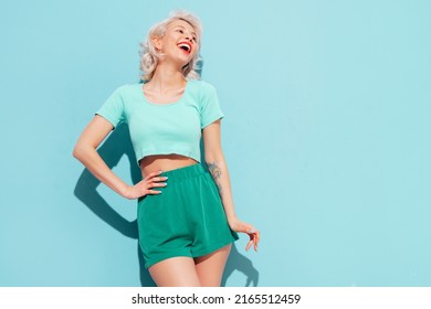 Young Beautiful Smiling Female In Trendy Summer Shorts And Top Clothes. Sexy Carefree Woman Posing Near Blue Wall In Studio. Positive Blond Model Having Fun And Going Crazy. Cheerful And Happy