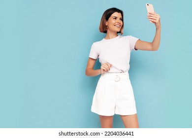 Young Beautiful Smiling Female In Trendy Summer Shorts Clothes. Carefree Woman Posing Near Blue Wall In Studio. Sexy Positive Model Having Fun Indoors. Cheerful And Happy. Taking Selfie