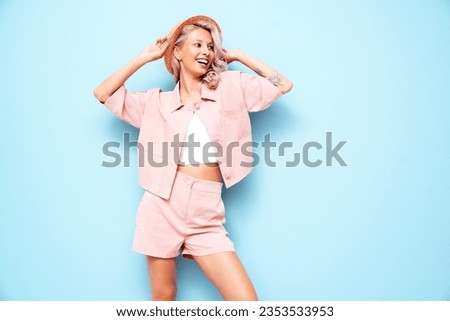 Young beautiful smiling female in trendy summer pink shorts and t-shirt. Carefree woman posing near blue wall in studio. Positive blond model having fun. Cheerful and happy. in hat