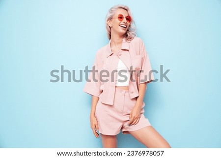 Young beautiful smiling female in trendy summer pink shorts and t-shirt. Carefree woman posing near blue wall in studio. Positive blond model having fun. Cheerful and happy