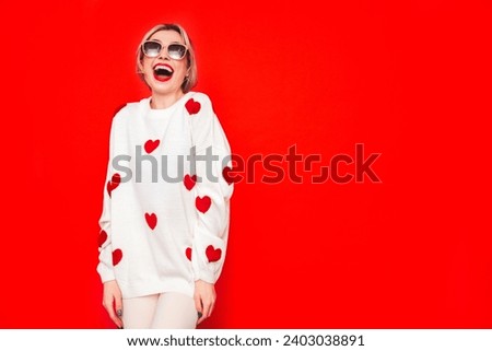 Young beautiful smiling female in trendy white sweater with hearts. Carefree woman posing near red wall in studio. Positive model having fun, going crazy. Cheerful and happy. Shocked and surprised 