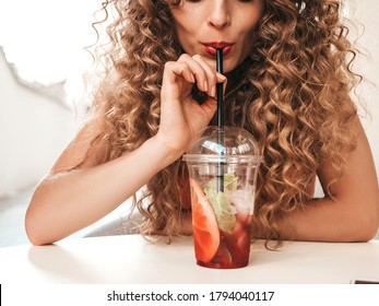 Young Beautiful Smiling Hipster Woman With Afro Curls Hairstyle. Woman In Summer Trendy Clothes Sitting In Veranda Cafe.Model Drinking Fresh Cocktail Smoothie Drink In Plastic Cup With Straw