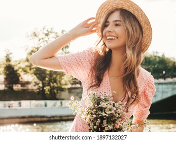 Young beautiful smiling hipster woman in trendy summer sundress. Sexy carefree woman posing on the street background in hat at sunset. Positive model outdoors on embankment. Holding flowers in hands
