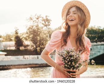 Young beautiful smiling hipster woman in trendy summer sundress. Sexy carefree woman posing on the street background in hat at sunset. Positive model outdoors on embankment. Holding flowers outdoors