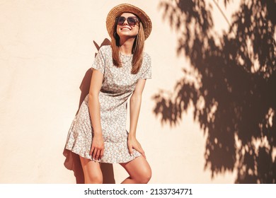 Young beautiful smiling hipster woman in trendy summer dress. Sexy carefree woman posing in the street near wall in hat at sunset. Positive model outdoors in sunglasses. Cheerful and happy