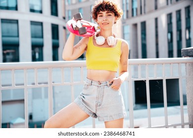Young beautiful smiling hipster woman in trendy summer clothes. Carefree woman with curls hairstyle, posing in the street. Positive model outdoors. Cheerful and happy. Holds penny skateboard - Powered by Shutterstock