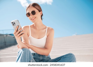 Young beautiful smiling hipster woman in trendy summer casual clothes. Carefree woman posing in the street. Positive model holds mobile phone, looks at cellphone screen, uses smartphone apps - Powered by Shutterstock