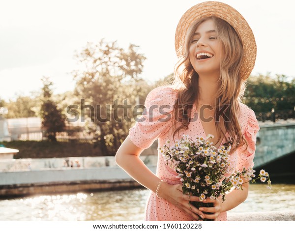 Young beautiful smiling hipster woman in trendy\
summer sundress. Sexy carefree woman posing on the street\
background in hat at sunset. Positive model outdoors on embankment.\
Holding flowers outdoors