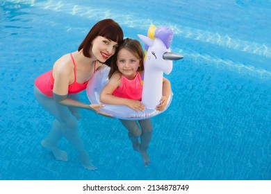 Young beautiful smiling mother teaching her daughter to swim in the pool with clear blue water. Daughter in unicorn lifebuoy with mom looking at camera while swimming in luxury pool. - Powered by Shutterstock