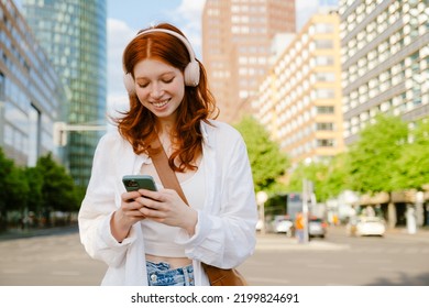 Young beautiful smiling redhead girl in headphones holding her phone both hands and looking on it, while standing in the city on the street - Powered by Shutterstock