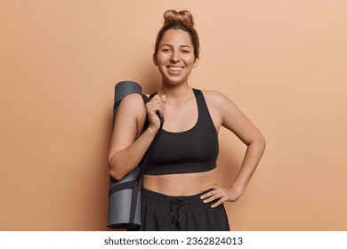 Young beautiful smiling sporty young woman with combed hair dressed in black tracksuit poses with rolled karemar returns home after training in gym has regular workout to keep fit and healthy.