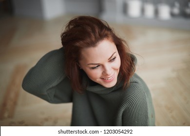 Young Beautiful Smiling Woman With Closed Eyes And Flowing Hair In Warm Green Sweater Enjoying Time At Home. Happy Life, Mental Health, Balance And Restore Concept. 