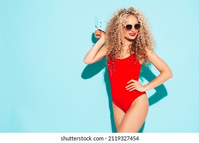 Young beautiful smiling woman posing near blue wall in studio.Sexy model in red swimwear bathing suit.Positive female with curls hairstyle. Holding penny skateboard.Happy and cheerful