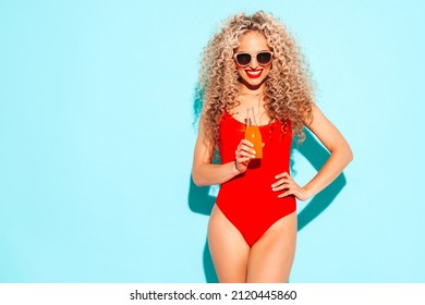 Young beautiful smiling woman posing near blue wall in studio.Sexy model in red swimwear bathing suit.Positive female with afro curls hairstyle. Happy and cheerful. Drinking lemonade from bottle
