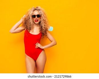 Young beautiful smiling woman posing near yellow wall in studio.Sexy model in red swimwear bathing suit.Positive female with curls hairstyle. Holding penny skateboard.Happy and cheerful