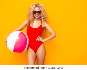 Young beautiful smiling woman posing near yellow wall in studio.Sexy model in red swimwear bathing suit.Positive female with curls hairstyle. Holding penny inflatable ball.Happy and cheerful