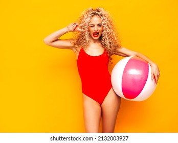 Young beautiful smiling woman posing near yellow wall in studio.Sexy model in red swimwear bathing suit.Positive female with curls hairstyle. Holding penny inflatable ball.Happy and cheerful