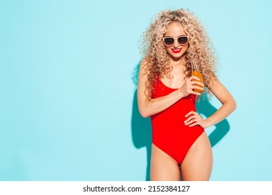 Young beautiful smiling woman posing near blue wall in studio.Sexy model in red swimwear bathing suit.Positive female with afro curls hairstyle. Happy and cheerful. Drinking lemonade from bottle